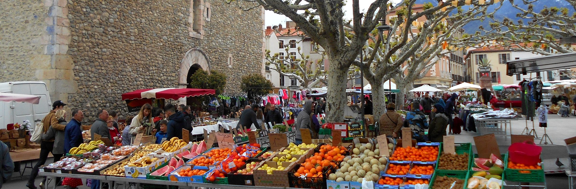 Openluchtmarkt onder de platanen van Prades: kleurrijke fruitkramen aan de voet van de kerk, bergen op de achtergrond